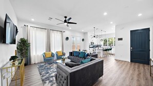 A modern living room with a sectional sofa, a coffee table, and a TV mounted on the wall. There is a dining area with a kitchen in the background. - Cozy Home Near MedCenter, Uptown, & Downtown (Houston)