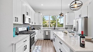 A modern kitchen with white cabinets, stainless steel appliances, and a large window offering a view of greenery. - Cozy Home Near MedCenter, Uptown, & Downtown (Houston)