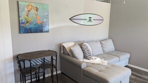 A living room with a grey sofa, a small wooden table, and two black metal chairs. A painting of two figures is on the wall, and a Chanel logo is also visible. - Cozy B- condo in charming Corpus Christi with WiFi, AC near Laguna Madre (Corpus Christi)