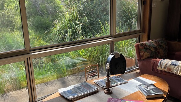A room with a large window, a wooden table with books and a lamp, a sofa with a floral cushion, and a vacuum cleaner. - One or Two rooms in house in O'Connor close to Civic, Dickson and Belconnen! (O'Connor)