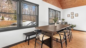 A dining area with a long wooden table, six chairs, and large windows offering a view of a snowy landscape. - Cozy 4-Bedroom Modern Mountaintop Retreat in Jewett  (Jewett)