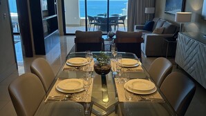 Un comedor con una mesa de vidrio preparada para seis personas, una lámpara de araña moderna y vistas al océano a través de grandes ventanales.