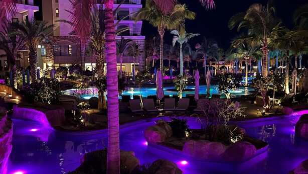 Zona de piscina en un resort durante la noche, con palmeras iluminadas y un edificio grande al fondo.