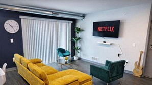 Una sala de estar con un sofá amarillo, un sillón azul verdoso y un televisor mostrando 'NETFLIX'.