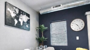 Un comedor con una mesa dispuesta para cuatro, sillas blancas, un mapa mundial en la pared, un reloj y una ventana con persianas.