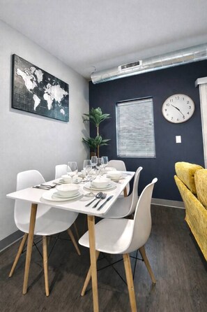 Un coin repas avec une table dressée pour quatre personnes, des chaises blanches, une carte du monde accrochée au mur, une horloge et une fenêtre avec des stores.
