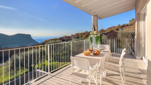 A balcony with a table set for dining, overlooking a scenic landscape with mountains and trees. - White Apartment with Terrace and Lake View (Costermano)