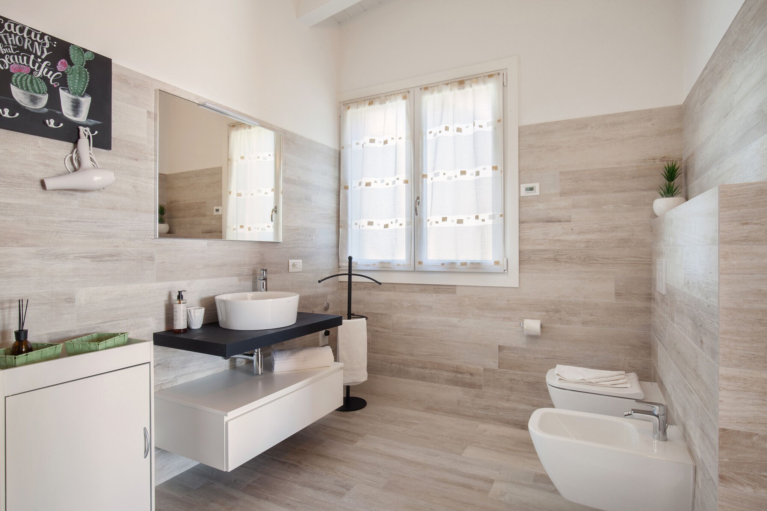 Une salle de bain moderne avec un lavabo, un meuble-lavabo et des toilettes, le tout de couleur blanche. On y trouve une fenêtre avec des rideaux, un miroir au-dessus du lavabo et une œuvre d’art sur le mur, dont le thème est celui des cactus.