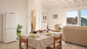 A dining area with a table set for two, a refrigerator, a sofa, and a window with a view of trees. - White Apartment with Terrace and Lake View (Costermano)