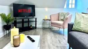 Ein modernes Wohnzimmer mit einem Flachbildfernseher, der 'NETFLIX' anzeigt, einem weißen Couchtisch, einem pinkfarbenen Sessel, einem grauen Sofa und einem großen Fenster mit Blick.