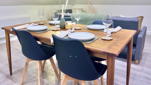 Ein Essbereich mit Holztisch, gedeckten Plätzen für vier Personen, dunklen Stühlen und gemütlicher Atmosphäre, beleuchtet durch eine Lampe und mit dekorativen Elementen.
