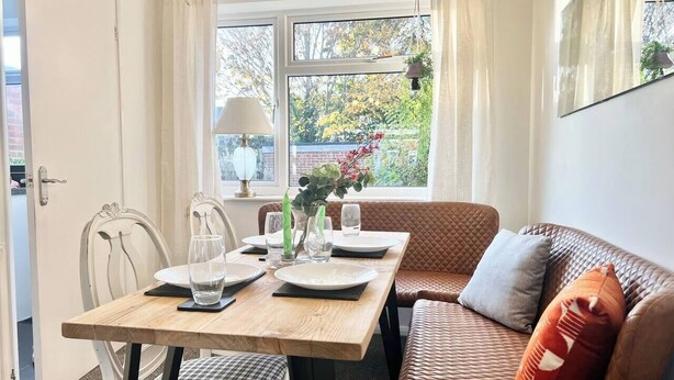 A dining area with a wooden table set for two, a brown cushioned sofa, and a window with curtains. - 3BR | Contractors | Parking, Garden (Saffron Walden)