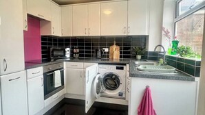 A compact kitchen with white cabinets, a built-in oven, a microwave, a washing machine, and a sink with a window above. - 3BR | Contractors | Parking, Garden (Saffron Walden)