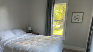 A neatly made bed with a white duvet, a bedside table with a lamp and a small plant, a window with a view of greenery, and a framed picture on the wall.
