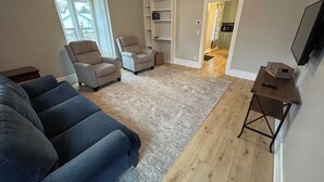 A living room with a large blue sofa, two armchairs, a wooden floor, and a TV on the wall.