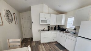 Una cocina compacta con armarios blancos, un microondas y un lavabo. Hay una mesa de comedor con sillas y una puerta con cerradura electrónica.