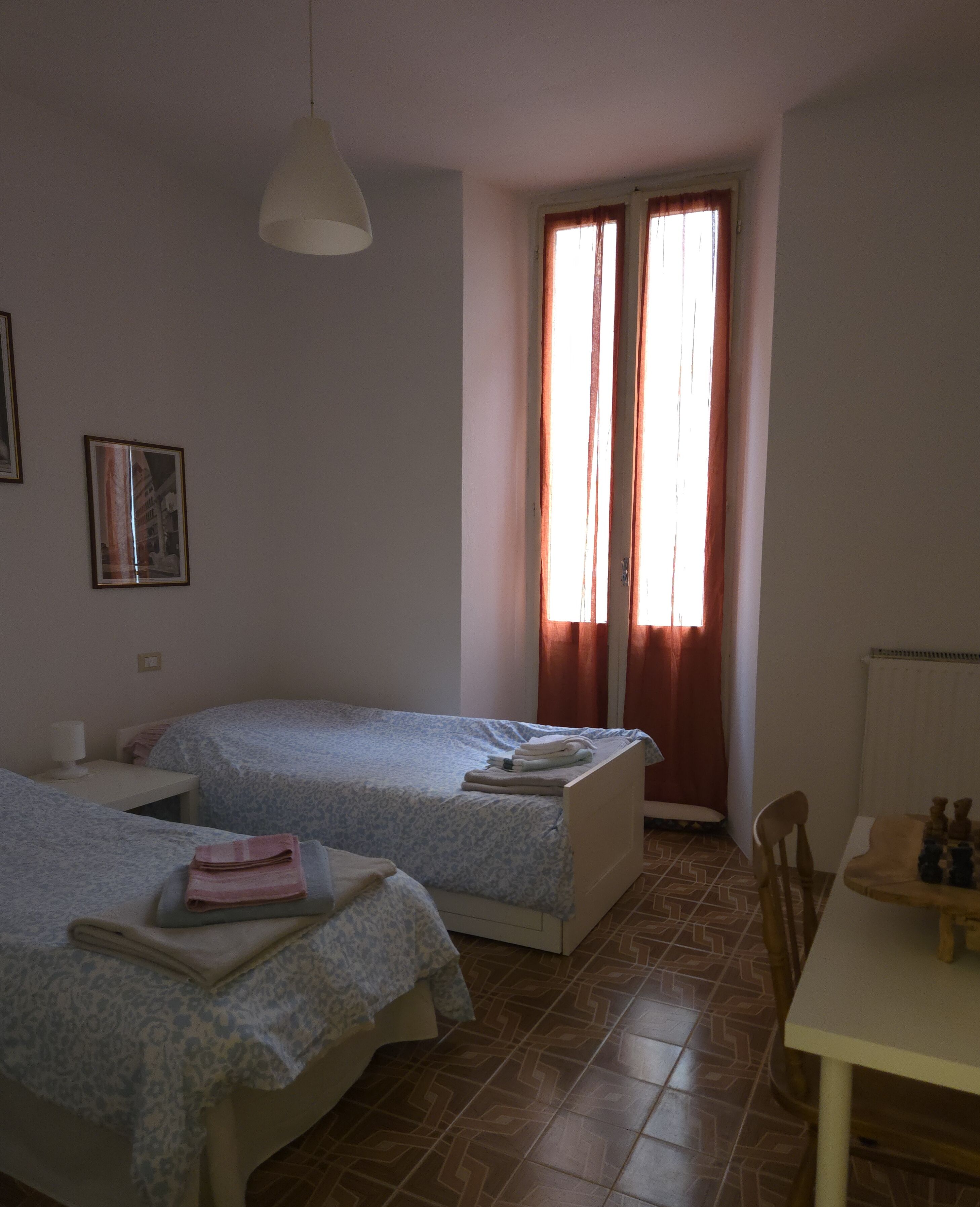 A room with two single beds, a table with a chess set, a hanging light, and a window with curtains.