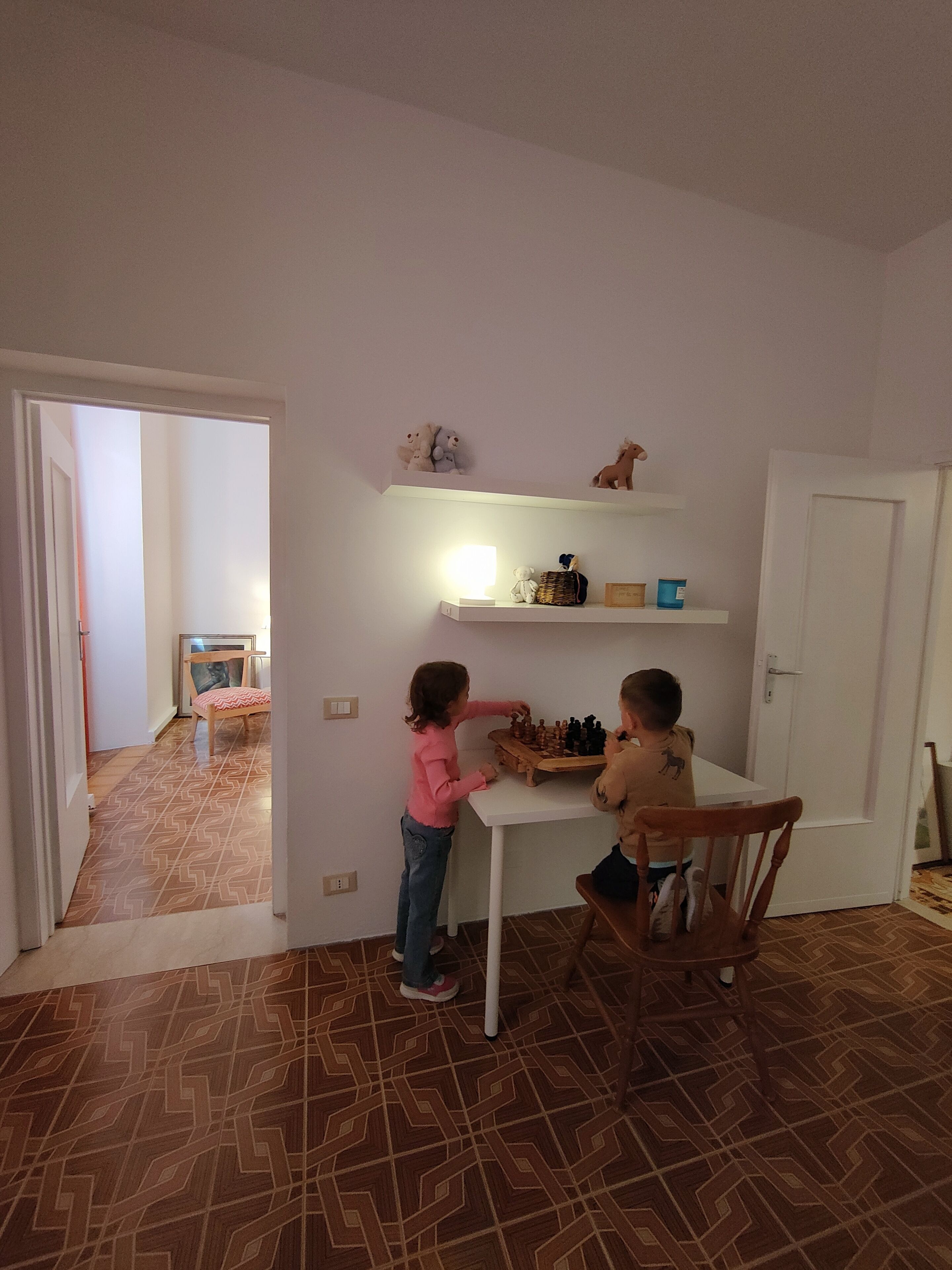 A room with a white table and chairs, a chessboard, and shelves with toys and a lamp.