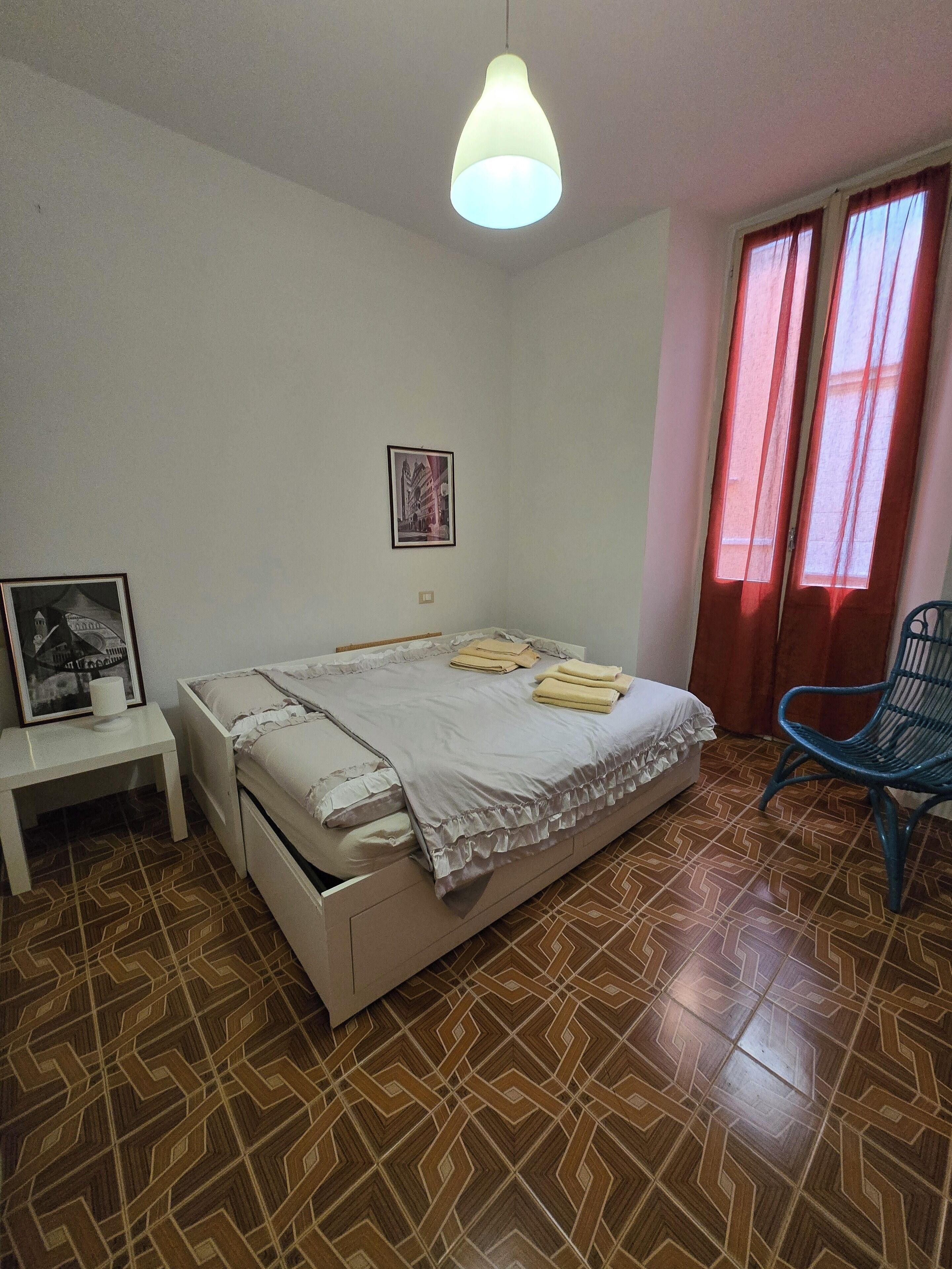 Ein Schlafzimmer mit Bett, Nachttisch, Stuhl, Pendelleuchte und einem fenster mit Vorhängen.