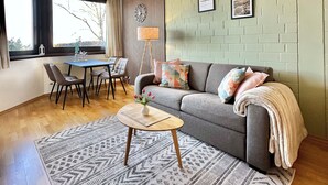 Ein modernes Wohnzimmer mit einem grauen Sofa, einem Holztisch, einem gemusterten Teppich und einem Essbereich mit Tisch und Stühlen.