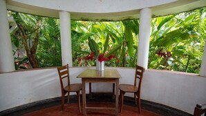A room with a table and two chairs, a vase with red flowers, and a view of lush green foliage through a window. - Thuru Sewana Villa (Unawatuna)