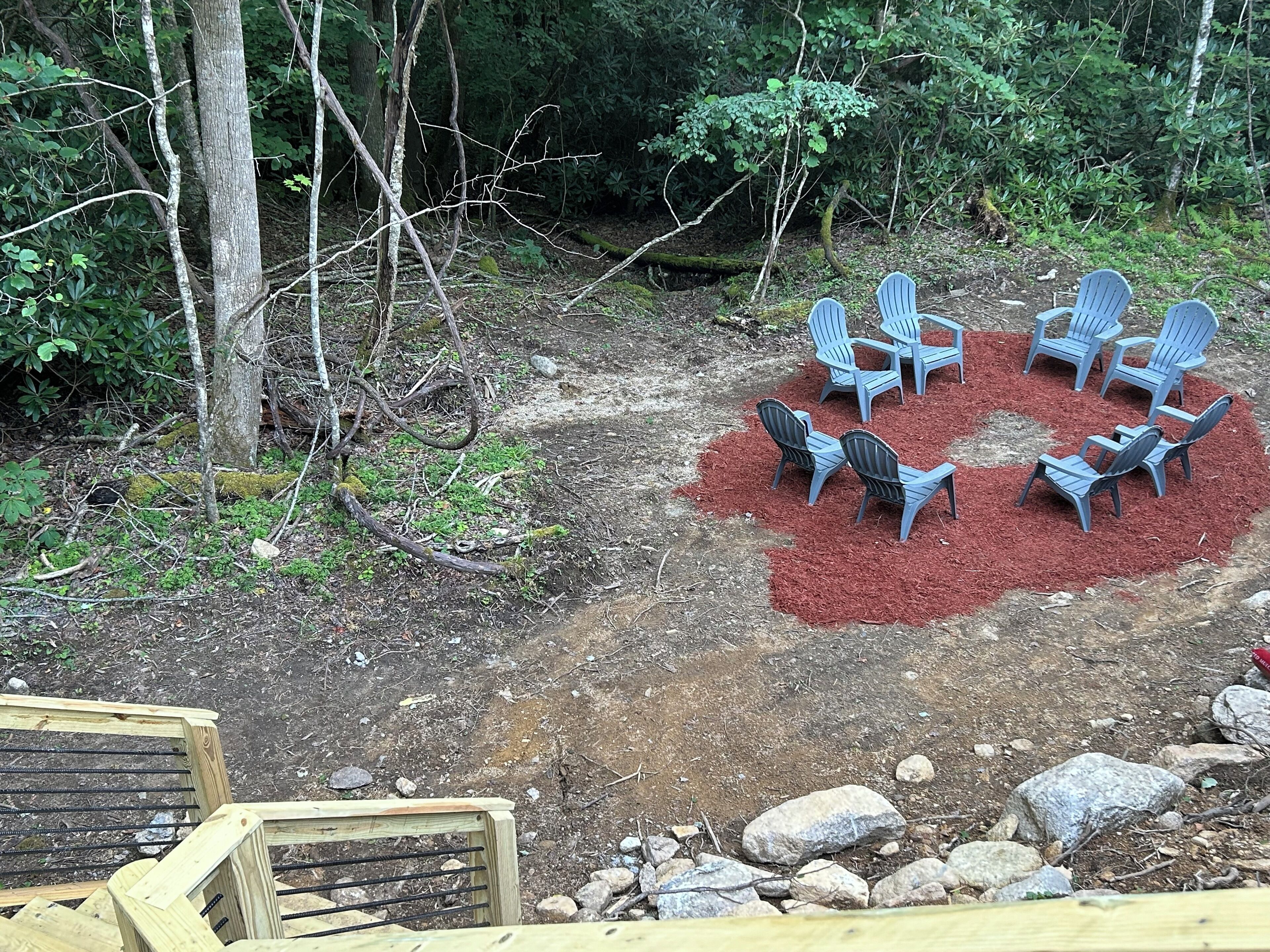 Ein runder Sitzbereich mit Stühlen und Tisch, umgeben von Holzmulch, inmitten einer bewaldeten Landschaft mit Bäumen und Steinen.