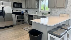A modern kitchen with stainless steel appliances, a large island with bar stools, and a view of the outdoors through a window. - Sea Level BSL 3BR 3BA Waterfront (Shoreline Park)