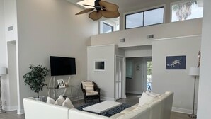 A spacious living room with a white sofa, a ceiling fan, a flat-screen TV, and a large window. - Sea Level BSL 3BR 3BA Waterfront (Shoreline Park)