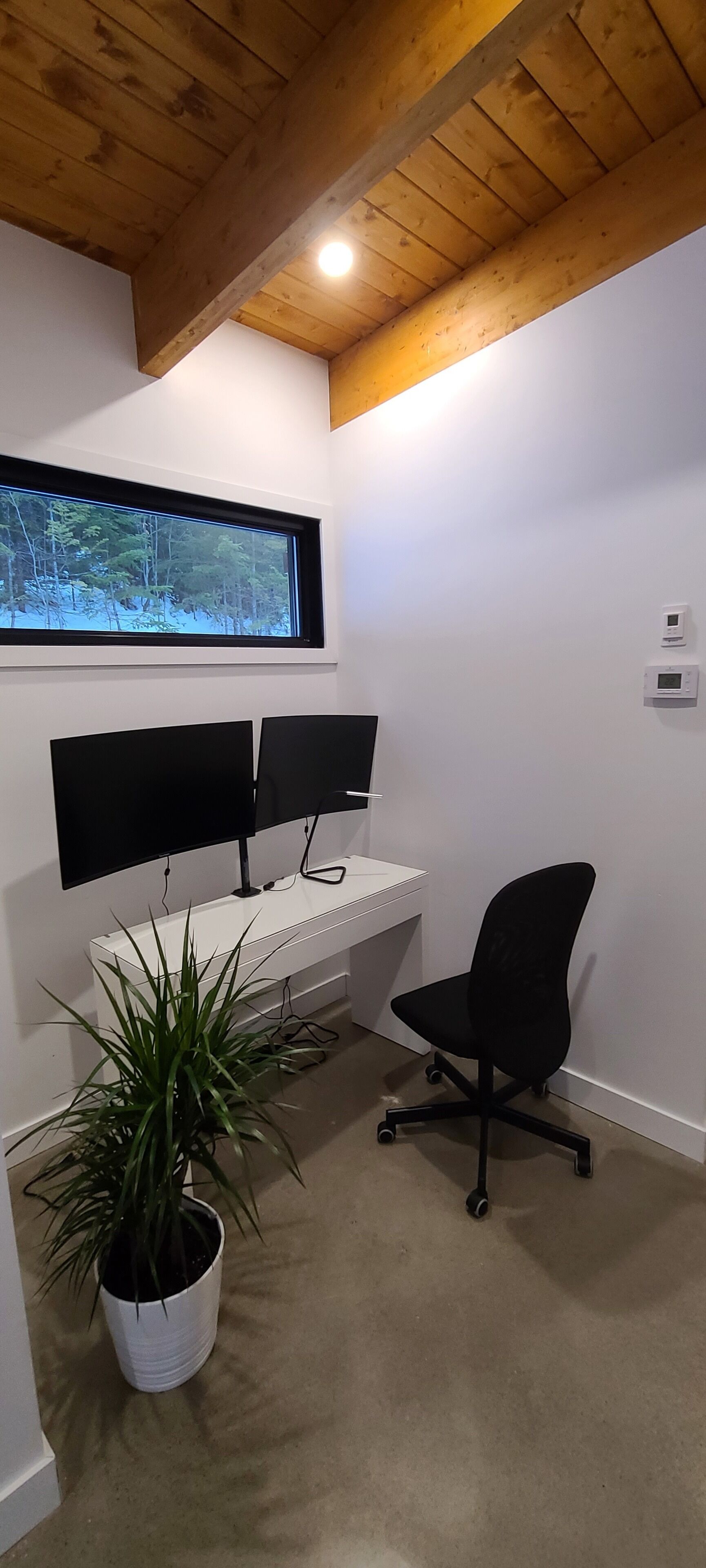 Ett rum med ett träpanelat tak, ett vitt skrivbord med två datorskärmar, en svart kontorsstol och en krukväxt.