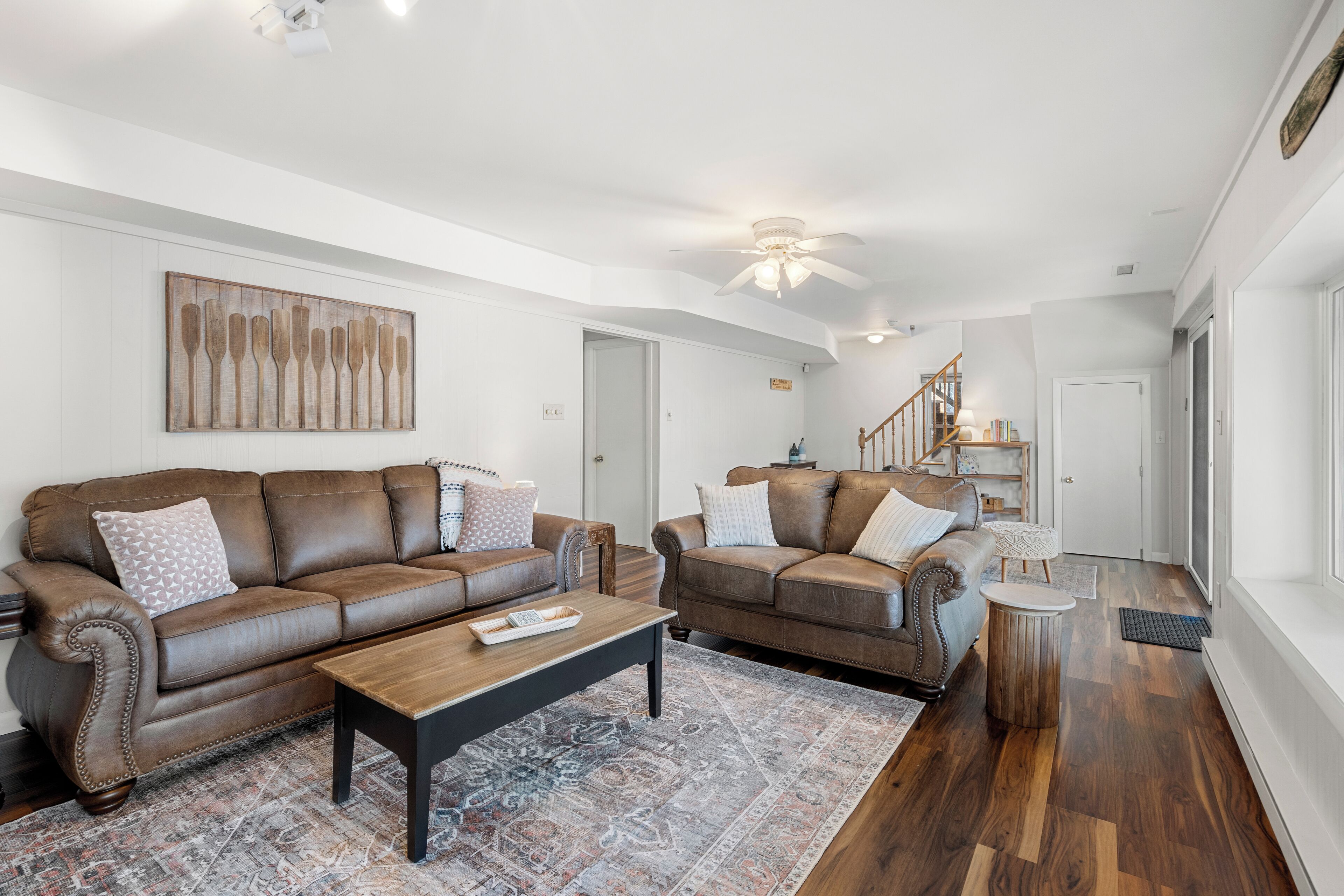 A spacious living room with two brown leather sofas, a wooden coffee table, and a patterned rug. A ceiling fan and a staircase are visible in the background.