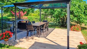 Un espacio comedor al aire libre cubierto, con mesa y sillas, rodeado de follaje y flores.