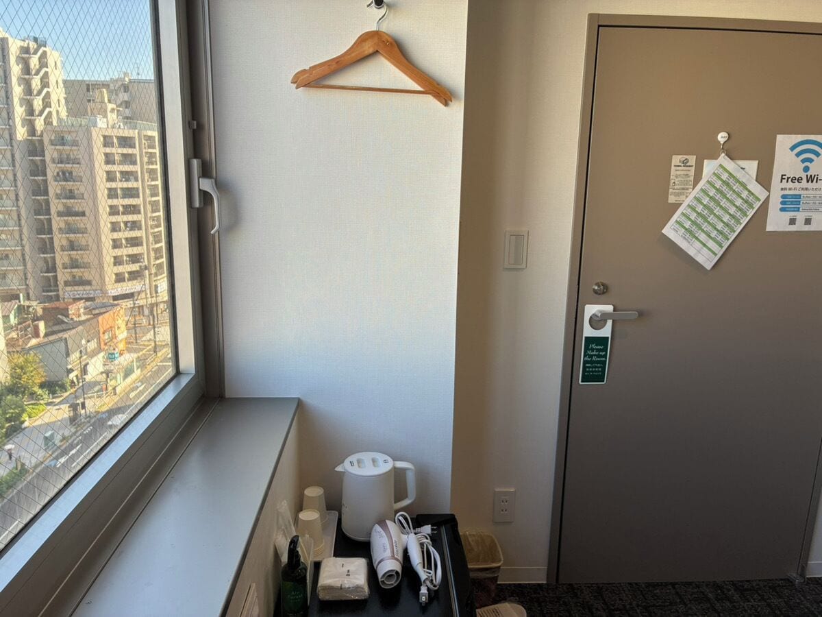 A hotel room with a window overlooking a cityscape, a wooden hanger, a door with a Wi-Fi sign, and a small table with toiletries.