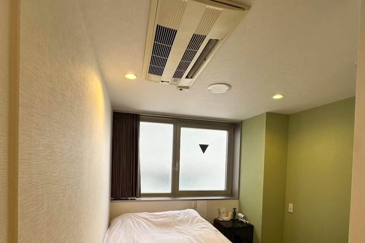 Habitación de hotel con cama, ventana con cortinas, mesita de noche con teléfono y un pequeño cubo de basura.