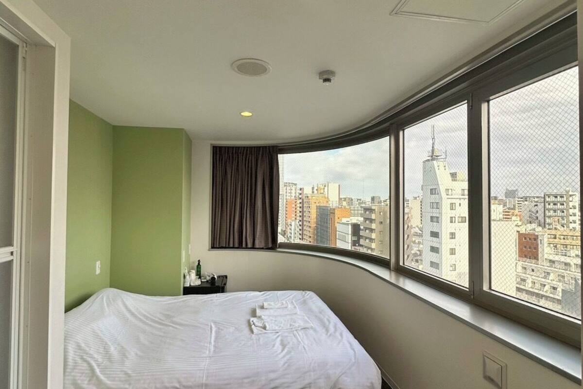 Una habitación de hotel con una cama, una ventana que ofrece vistas a edificios, una cortina y una lámpara en el techo.