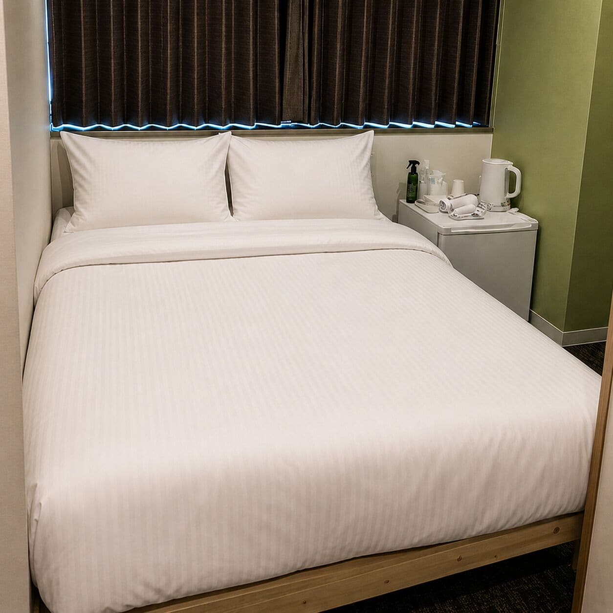 A neatly made bed with white linens, a bedside table with a phone, a bottle, and a cup.