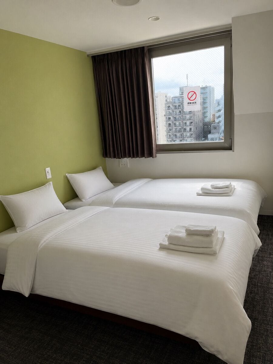 A hotel room with two beds, a window with a view of buildings, and a no-smoking sign.