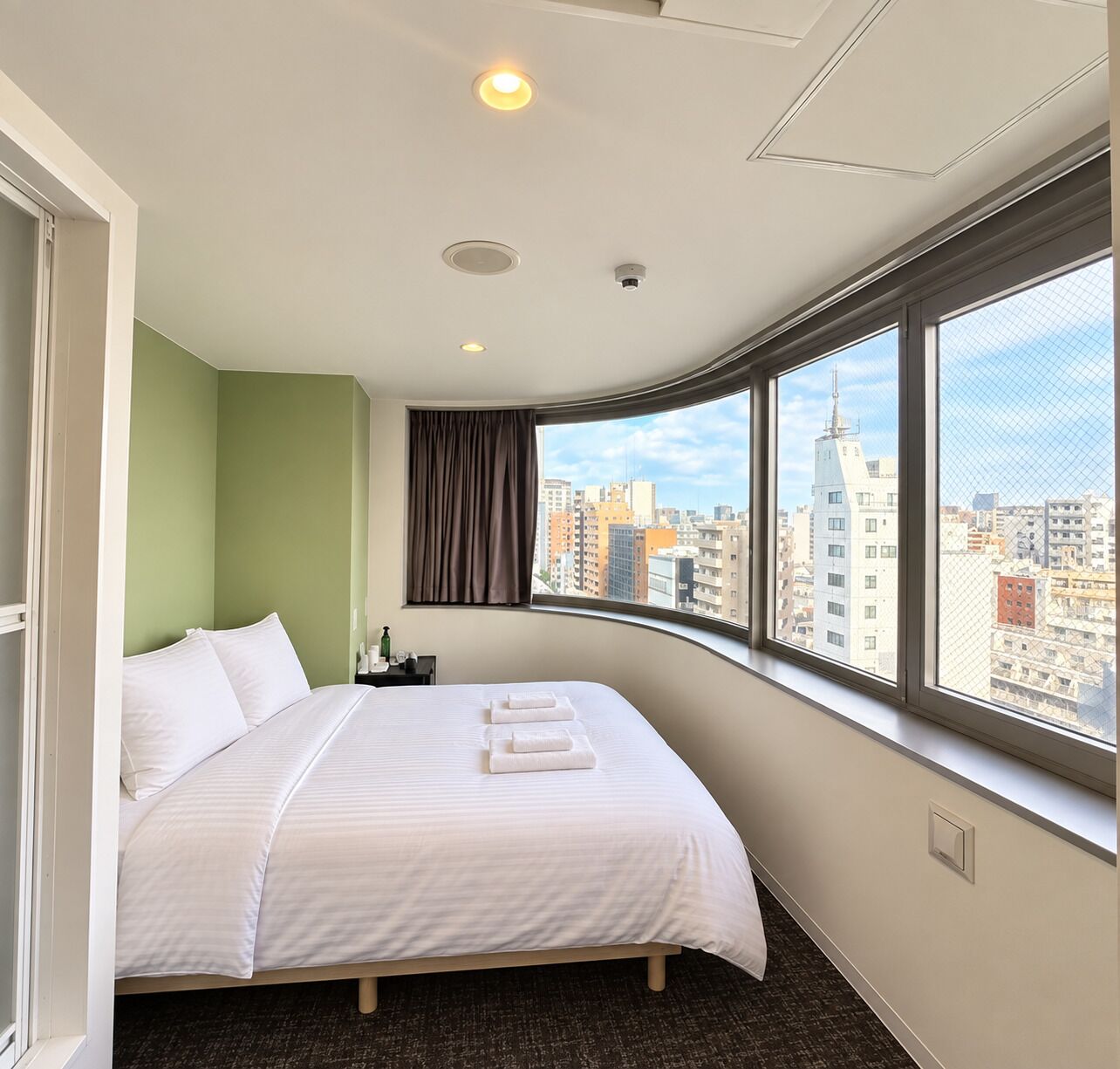 A hotel room with a bed, a nightstand, a window with a city view, and a ceiling with recessed lighting.