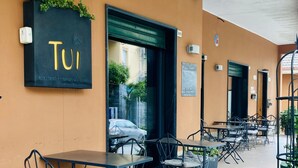 An outdoor café area with tables and chairs, a sign reading "TUI," and a door leading inside. - Tuibeds (Arenzano)