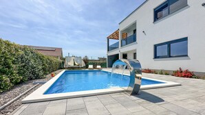 A modern outdoor pool area with a water feature, a bed with a canopy, and a two-story building with large windows.