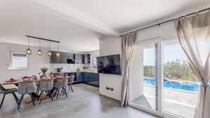 A modern kitchen with a dining area, a large TV, and a view of a swimming pool.