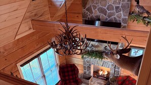 A multi-level cabin with a stone fireplace, a deer head mounted on the wall, and a chandelier with antlers.