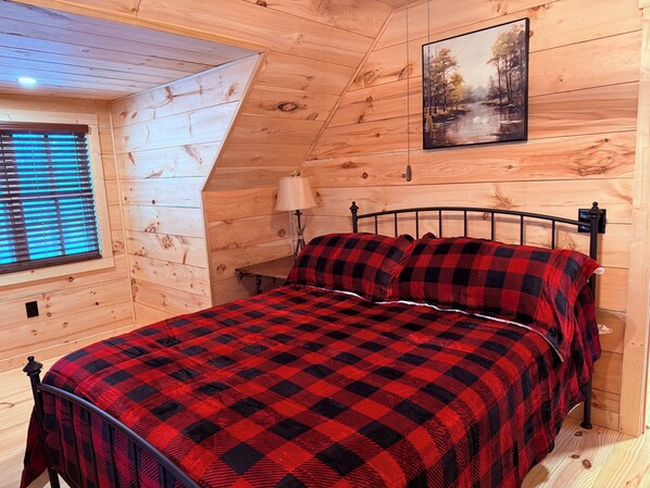 A cozy wooden cabin bedroom with a bed, a nightstand, a lamp, and a window with blinds.