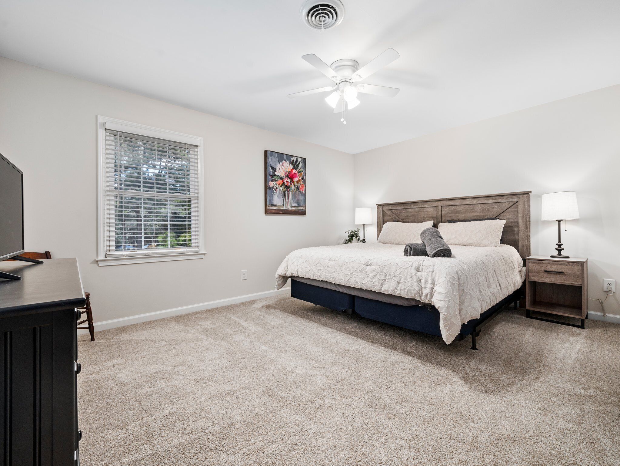 A bedroom with a bed, bedside tables, a ceiling fan, a window with blinds, and a painting on the wall.