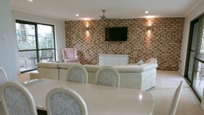 A dining area with a white table and chairs, a sofa, and a TV mounted on a brick-patterned wall. - Spacious, comfortable, Pet Friendly 3-Bed House minutes from the Big Pineapple  (Woombye)