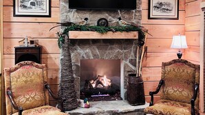 A cozy living room with a stone fireplace, two ornate chairs, a television mounted above the fireplace, and framed pictures on the walls.