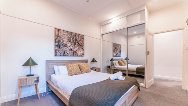 A bedroom with a bed, bedside lamps, a large abstract painting, a wardrobe, and a mirror. - 3BR Glen Iris Gem – Sleeps 7, Near Train & Freeway with WiFi and AC (Glen Iris)