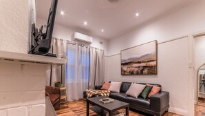 A living room with a sofa, a coffee table, a TV on a stand, a painting on the wall, and a window with curtains. - 3BR Glen Iris Gem – Sleeps 7, Near Train & Freeway with WiFi and AC (Glen Iris)