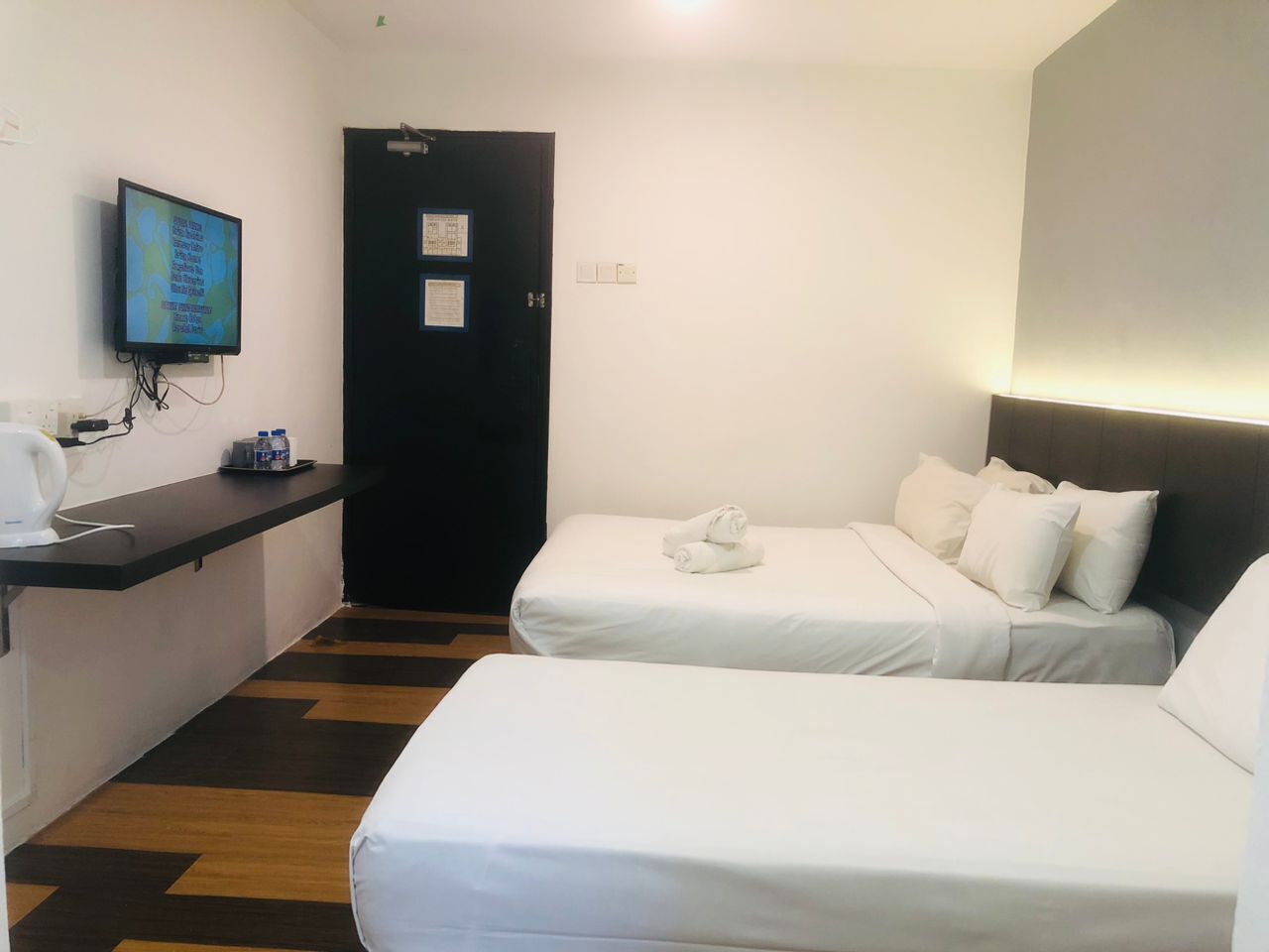 A hotel room with two beds, a desk with a computer monitor, and a door with a notice.