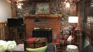 A cozy living room with a fireplace, a television on a wooden stand, a plaid armchair, and a small table with a vase of flowers. - 1st Floor - 1 mile from San Moritz Lodge (Crestline)