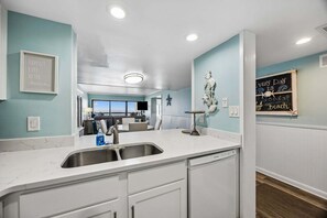 A kitchen with a white countertop, a sink, and a view into the dining area. There are wall decorations including a framed quote and a chalkboard with a message. - "Seventh Heaven" Beachfront Condo Near The Ritz! 2 Pools, Luxury Property (Fernandina Beach)